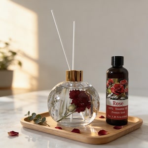 May include: A clear glass diffuser with a gold cap and white reeds, containing a red flower, sits on a wooden tray next to a brown bottle labeled "Rose 100% Essential Oil". The tray also holds rose petals and greenery.