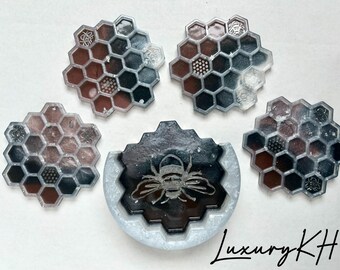 Puede incluir: Un conjunto de posavasos y una bandeja decorativa de resina. Los posavasos y la bandeja presentan un diseño de panal con tonos marrones, negros y grises. La bandeja tiene un diseño de abeja. El texto "Luxury KH" es visible.