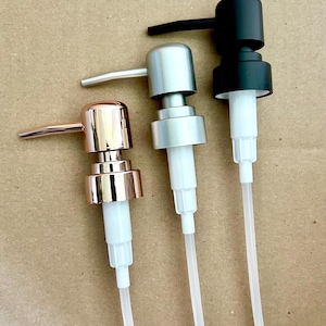 May include: Three different soap dispenser pumps. One is rose gold, one is silver, and the other is black. All have white plastic tubes. The pumps are on a light brown surface.