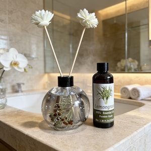 Op de afbeelding: Een heldere, bolvormige diffuserfles met een zwarte dop en twee witte bloemvormige rietjes. Een kleinere bruine fles citroengras etherische olie staat ernaast. Het etiket luidt "Lemongrass 100% Essential Oil Premium Grade Net 100ml".