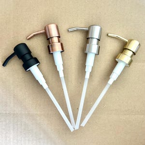 May include: Four soap dispenser pumps in black, copper, silver, and gold finishes. Each pump has a white plastic tube and a curved spout. The pumps are arranged diagonally against a neutral background.