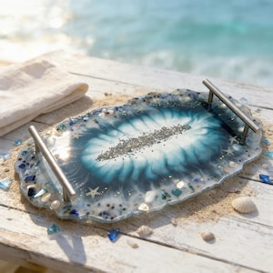 Pode incluir: Uma bandeja decorativa de resina com tema costeiro. A bandeja apresenta um design azul e branco com conchas, estrelas do mar e pedaços de vidro embutidos. Possui alças prateadas.