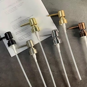 May include: Assortment of soap or lotion dispenser pumps in various metallic finishes: gold, silver, bronze, and black. Each pump has a long white tube. The pumps are displayed on a light-colored surface with text that reads "CHAMPAGNE ROSES" and "LOVE AND HOPE".