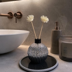 May include: A glass reed diffuser with a round, ribbed body that transitions from clear at the top to dark gray at the base. Two cream-colored artificial flowers are attached to wooden reeds inserted into the black neck of the bottle, which sits on a circular black and gray tray.