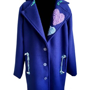 Könnte beinhalten: Ein marineblauer Mantel mit einem großen herzförmigen Design am Kragen. Das Herz ist aus bunten Perlen und Pailletten gefertigt. Der Mantel hat zwei Taschen auf der Vorderseite, jede mit einem Perlenmuster.