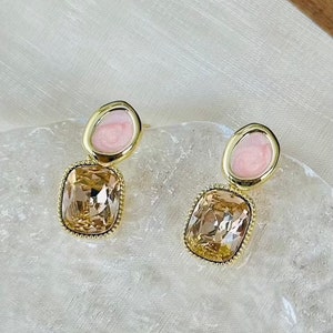 May include: A pair of gold earrings with a pink oval-shaped stone and a larger, square-shaped, light brown stone.