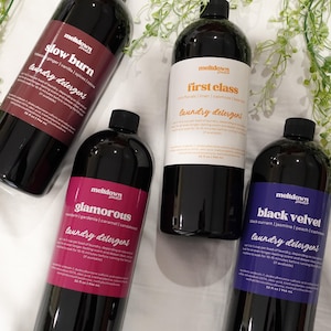 May include: Four black bottles of laundry detergent with different labels. The labels read "Slow Burn", "First Class", "Glamorous", and "Black Velvet". Each bottle is 946 ml.