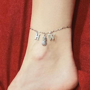 May include: Silver-toned anklet with the letters "H" and "W" and a pineapple charm. The delicate chain is worn on an ankle against a red background. The anklet is a piece of jewelry.