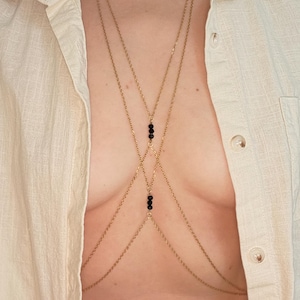 May include: A gold-colored body chain with black bead accents. The chain drapes across the chest in a layered design. The jewelry is worn over a light-colored button-down shirt.