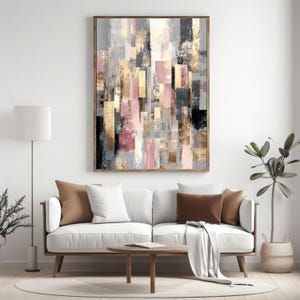 May include: Abstract art print in a wooden frame. The artwork features a collage of rectangular shapes in shades of gray, pink, gold, and black. The painting hangs above a white sofa with brown and white pillows, a round wooden coffee table, and a floor lamp.