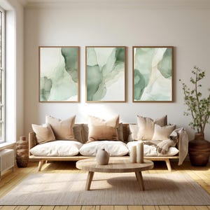 Peut inclure: Trois impressions d'art abstrait encadrées avec des motifs de type aquarelle vert et blanc. Les impressions sont accrochées à un mur blanc au-dessus d'un canapé brun clair avec des coussins beiges et une table basse en bois.