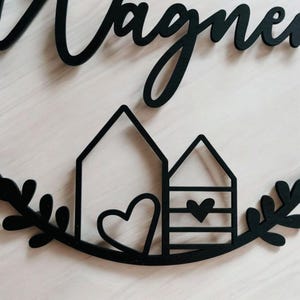 Puede incluir: Un letrero de madera negro con la palabra "Wagner" escrita en cursiva. El letrero presenta dos peque&ntilde;as casas con corazones y una corona de hojas.