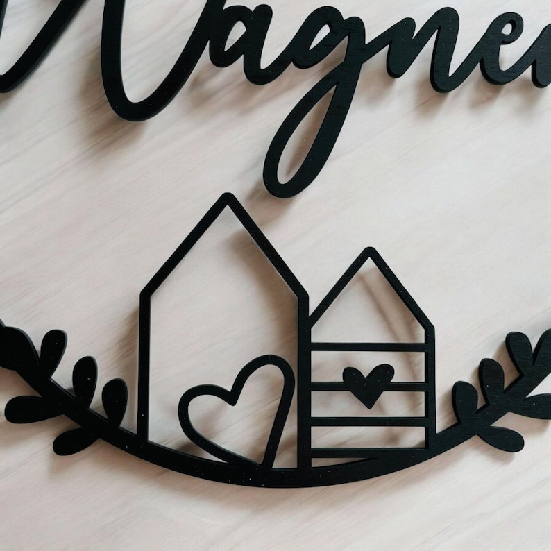 Puede incluir: Un letrero de madera negro con la palabra "Wagner" escrita en cursiva. El letrero presenta dos peque&ntilde;as casas con corazones y una corona de hojas.