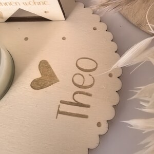 May include: A wooden round plaque with a scalloped edge, featuring a heart and the name "Theo" engraved in a light brown color. The plaque is decorated with white feathers.