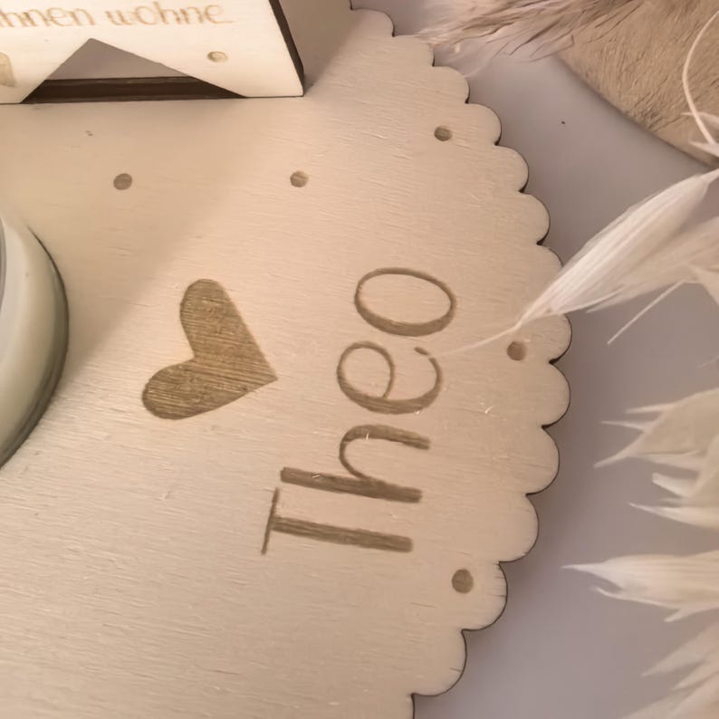 May include: A wooden round plaque with a scalloped edge, featuring a heart and the name "Theo" engraved in a light brown color. The plaque is decorated with white feathers.