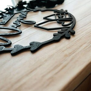 Puede incluir: Un letrero de madera negro con las palabras "Family Forever" en una fuente cursiva, rodeado de una corona de hojas y corazones. El letrero est&aacute; sobre una superficie de madera clara.