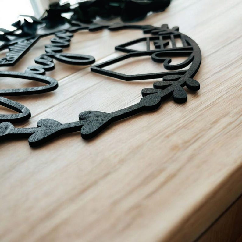 Puede incluir: Un letrero de madera negro con las palabras "Family Forever" en una fuente cursiva, rodeado de una corona de hojas y corazones. El letrero est&aacute; sobre una superficie de madera clara.