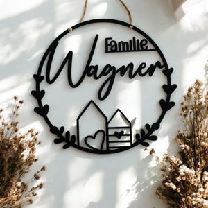 Puede incluir: Un letrero redondo de madera negro con el texto "Familie Wagner" en una fuente cursiva. El letrero tiene un dise&ntilde;o de corona con dos peque&ntilde;as casas y corazones.