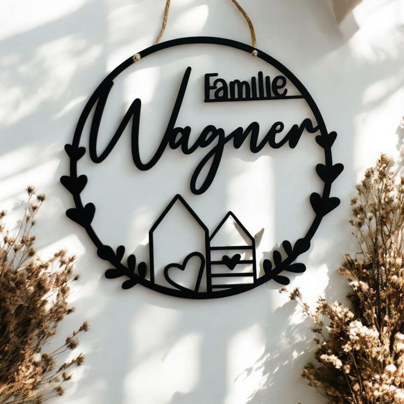 Puede incluir: Un letrero redondo de madera negro con el texto "Familie Wagner" en una fuente cursiva. El letrero tiene un dise&ntilde;o de corona con dos peque&ntilde;as casas y corazones.