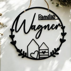 Puede incluir: Un letrero redondo de madera negro con el texto "Familie Wagner" en una fuente cursiva. El letrero est&aacute; decorado con una corona de hojas y dos peque&ntilde;as casas con corazones.