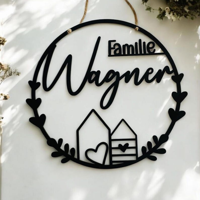 Puede incluir: Un letrero redondo de madera negro con el texto "Familie Wagner" en una fuente cursiva. El letrero est&aacute; decorado con una corona de hojas y dos peque&ntilde;as casas con corazones.
