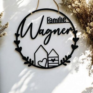 Puede incluir: Un letrero redondo de madera negro con el texto "Familie Wagner" en una fuente cursiva. El letrero tiene un dise&ntilde;o de corona con peque&ntilde;os corazones y dos peque&ntilde;as casas con corazones en la parte inferior.