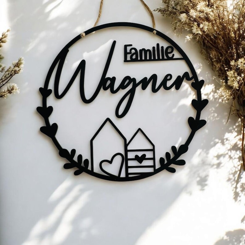 Puede incluir: Un letrero redondo de madera negro con el texto "Familie Wagner" en una fuente cursiva. El letrero tiene un dise&ntilde;o de corona con peque&ntilde;os corazones y dos peque&ntilde;as casas con corazones en la parte inferior.