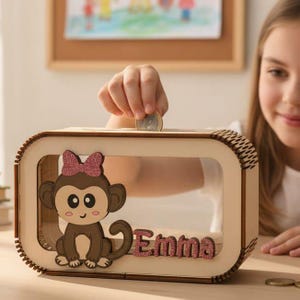 Könnte beinhalten: Eine Holzspardose mit einer transparenten Frontplatte. Die Box zeigt einen Cartoon-Affen mit einer rosa Schleife und dem Namen "Emma" in rosa Glitzerbuchstaben. Eine Münze wird in den Schlitz oben eingefügt. Die Box ist rechteckig mit abgerundeten Ecken.