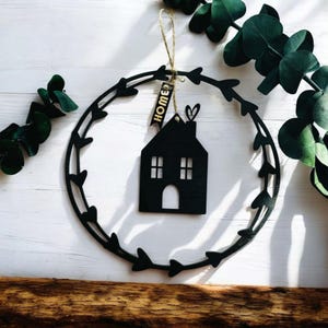 Può includere: Silhouette nera di una casa in legno con un'etichetta dorata "Home" appesa a una ghirlanda nera con foglie a forma di cuore.
