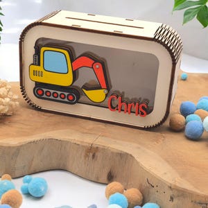Könnte beinhalten: Holz-Sparschwein in Form eines Baggers mit einem transparenten Plastikfenster, um die Münzen im Inneren zu sehen. Der Bagger ist gelb mit roten Armen und einer schwarzen Raupe. Der Name "Chris" steht auf der Vorderseite des Sparschweins.