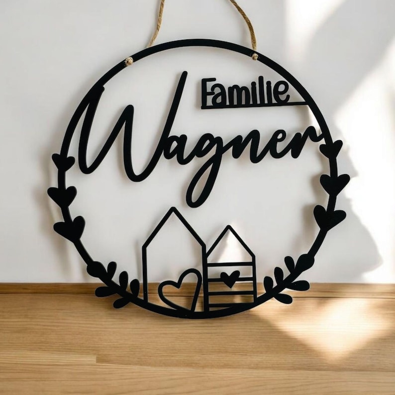 Puede incluir: Un letrero redondo de madera negro con el texto "Familie Wagner" en una fuente cursiva. El letrero tiene un dise&ntilde;o de corona con dos peque&ntilde;as casas y un coraz&oacute;n en el centro.