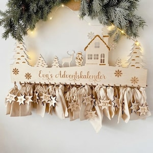 Könnte beinhalten: Ein hölzerner Adventskalender zum Aufhängen mit einer lasergeschnittenen Winterlandschaft aus Haus, Schneemann, Hirsch und Tannen. Der Schriftzug "Eva's Adventskalender" ist mittig eingraviert, ergänzt durch vierundzwanzig kleine Stoffbeutel.