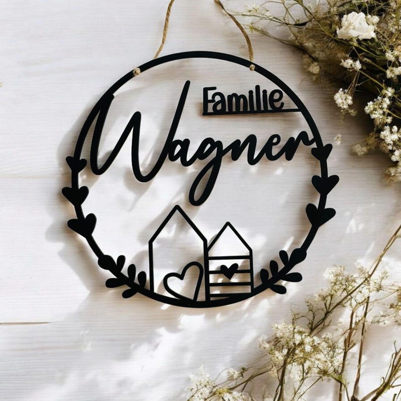 Puede incluir: Un letrero de madera negro con el texto "Familie Wagner" en una fuente cursiva. El letrero tiene la forma de un c&iacute;rculo con un dise&ntilde;o de corona alrededor del borde. Dos peque&ntilde;as casas con corazones est&aacute;n en el centro del c&iacute;rculo.