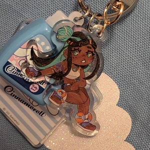 May include: A keychain featuring a cartoon character with dark skin, teal hair, and a white top. The character is on a clear acrylic charm. A light blue charm with the text "Cinnamoroll" is also attached. The keychain has a silver clasp.