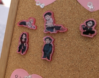 May include: Five colorful character pins on a corkboard. The pins are made of clear plastic with a pink backing. The characters are drawn in a cartoon style and are all wearing different outfits.