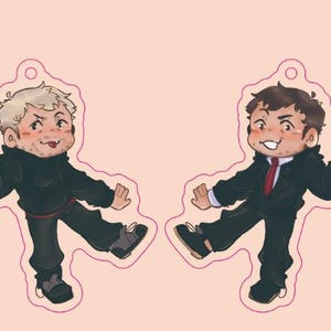 May include: Two cartoon character keychains. One keychain features a character with white hair and a black outfit, holding a golden object. The other keychain features a character with brown hair, a black suit, and a red tie, also holding a golden object.