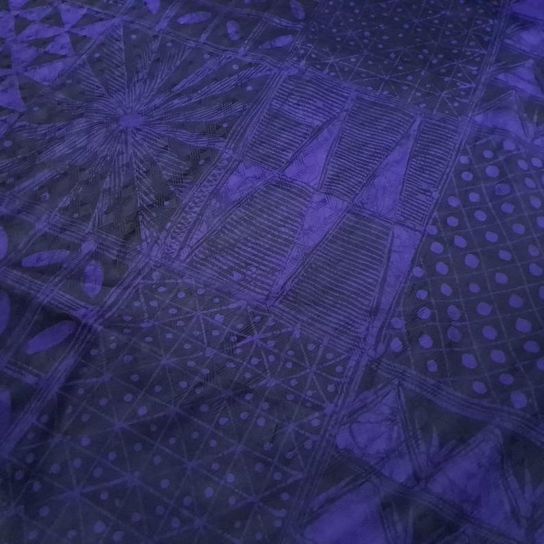 May include: A close-up of a textile with a patchwork design in shades of purple and dark blue. The fabric features geometric patterns, including squares, triangles, and circles, with intricate line work and dot patterns. The overall effect is a textured, artistic design.