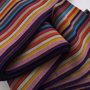 May include: Multicolored striped fabric ribbon with a purple backing. The ribbon is folded and stacked, showing the different colors and patterns.