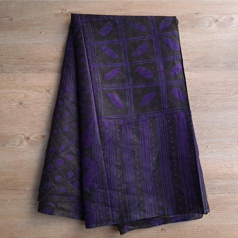 May include: Folded fabric with a repeating pattern of purple leaf shapes and grid lines on a black background. The fabric is draped over a wooden surface, showcasing the design and texture.
