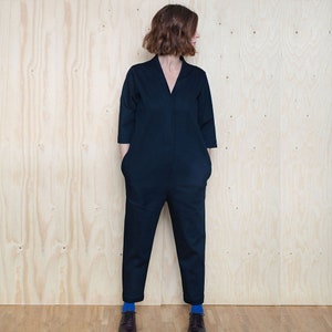 May include: A dark blue jumpsuit with a v-neck and long sleeves. The jumpsuit has pockets on the front and is worn by a person with short brown hair. The person is standing in front of a wood panel background.