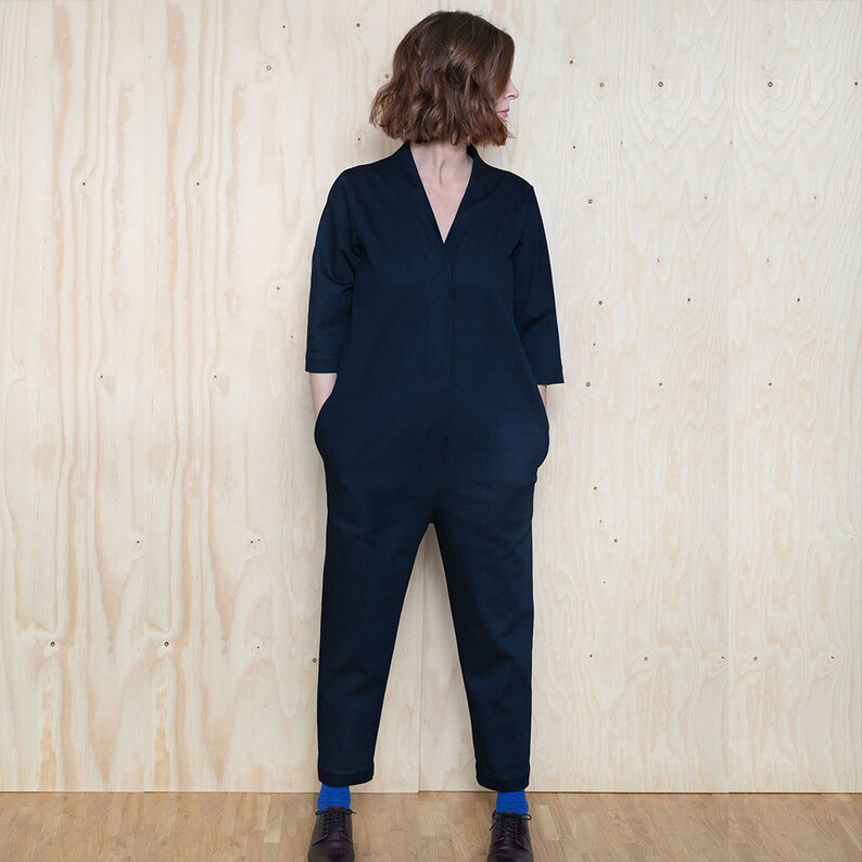 May include: A dark blue jumpsuit with a v-neck and long sleeves. The jumpsuit has pockets on the front and is worn by a person with short brown hair. The person is standing in front of a wood panel background.