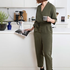 May include: A woman in a green jumpsuit stands in a kitchen, holding a cup of coffee and reading a magazine. She is wearing black ankle boots.