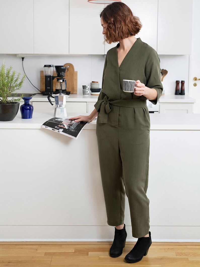 May include: A woman in a green jumpsuit stands in a kitchen, holding a cup of coffee and reading a magazine. She is wearing black ankle boots.