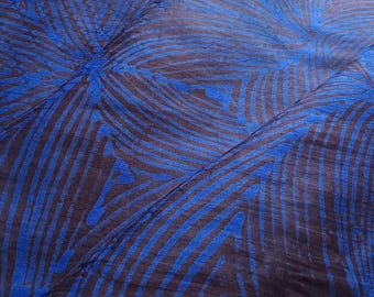 May include: Close-up of a fabric with a blue and brown abstract pattern. The design features curved, wave-like lines in a vibrant blue color against a darker brown background. The fabric is folded, showing the texture and drape of the material.