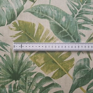 May include: A close-up of a beige fabric with a green tropical leaf print. A ruler is placed on the fabric, showing the scale in inches.