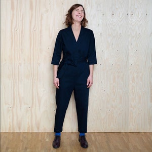 May include: A woman wearing a dark blue jumpsuit with a tie belt. She is standing in front of a wood panel background and smiling.