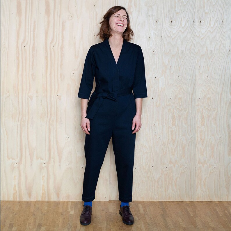 May include: A woman wearing a dark blue jumpsuit with a tie belt. She is standing in front of a wood panel background and smiling.