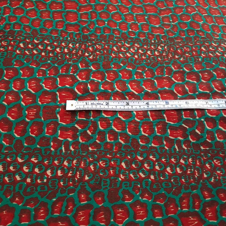 May include: Red and green fabric with a repeating pattern of small, rounded shapes. The fabric is measured with a ruler, showing 7 inches.