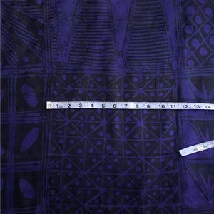 May include: Close-up of a fabric with a geometric pattern in black and purple. The design includes squares, triangles, and oval shapes. A measuring tape is placed across the fabric, showing inches.