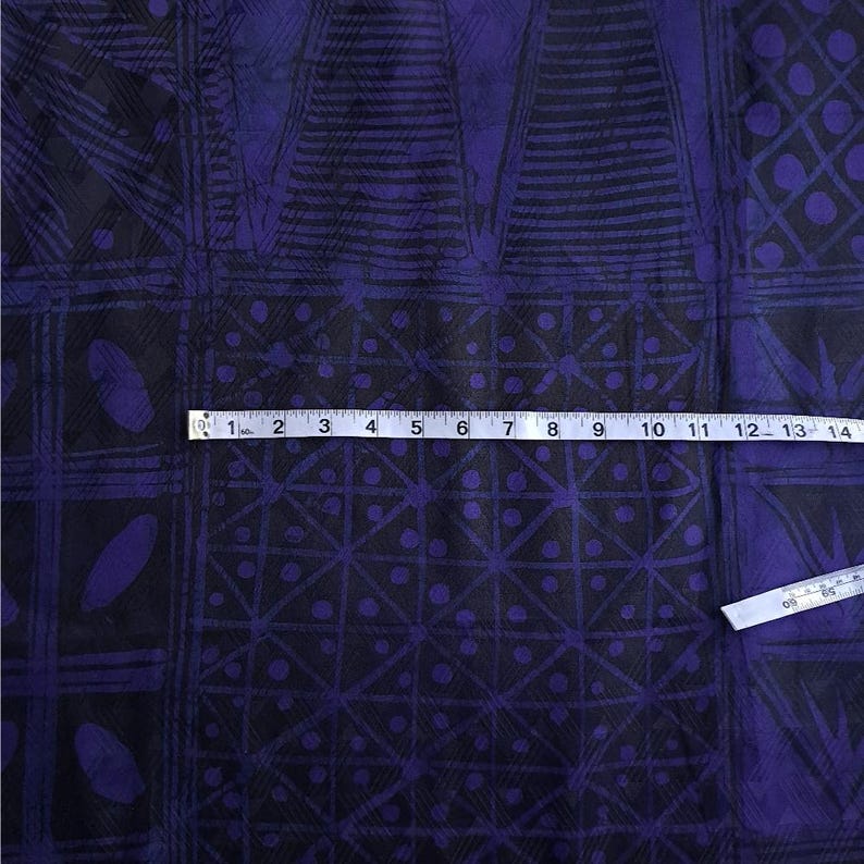 May include: Close-up of a fabric with a geometric pattern in black and purple. The design includes squares, triangles, and oval shapes. A measuring tape is placed across the fabric, showing inches.